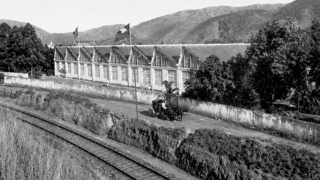 Fotografia antiga em ângulo elevado mostrando trilhos de trem em primeiro plano e o longo galpão fabril com telhado serrilhado.