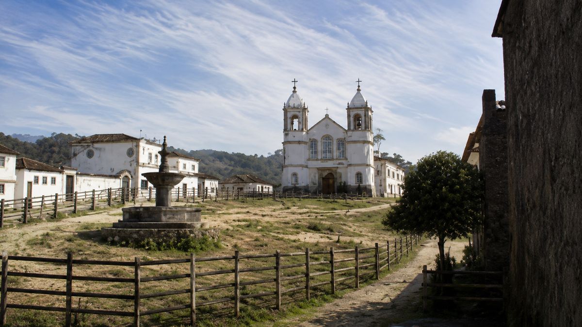 Fotografia da antiga vila de São João Marcos mostrando uma igreja colonial branca com duas torres sineiras ao fundo, uma fonte de pedra em primeiro plano e cercas de madeira delimitando caminhos em um terreno gramado sob céu azul com nuvens alongadas.