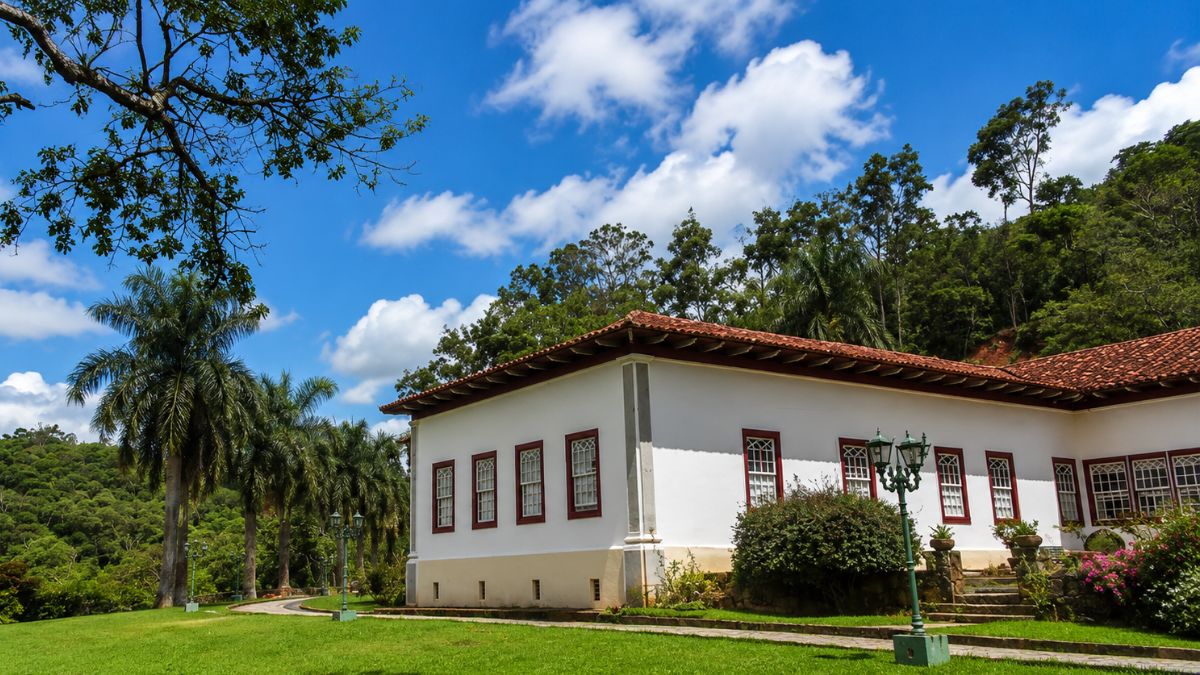 Fotografia da fachada lateral de um casarão colonial branco com janelas de molduras vermelhas, cercado por um amplo gramado verde, palmeiras imperiais e árvores sob um céu azul com nuvens.
