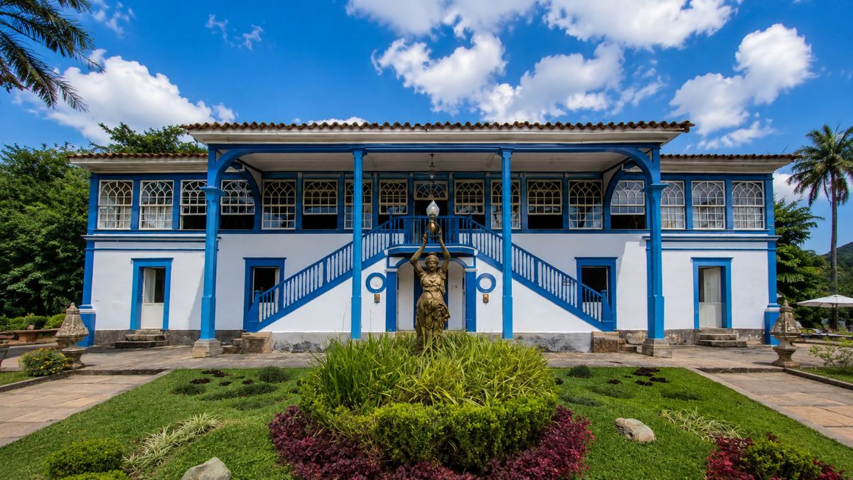Fotografia da fachada de um casarão colonial de dois andares nas cores branca e azul com escadaria simétrica e estátua central em um jardim.