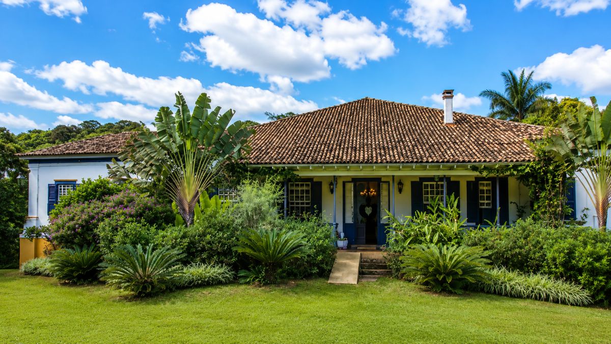 Fotografia da fachada de um casarão colonial de um pavimento, com telhado de barro, janelas azuis, cercado por um jardim verdejante com palmeiras e gramado sob céu azul com nuvens brancas.