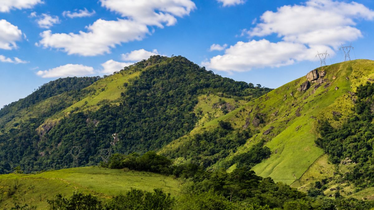 Fotografia das colinas verdes e montanhas cobertas por vegetação nativa do Vale do Café sob um céu azul com nuvens brancas, destacando duas torres de transmissão de energia elétrica no topo de um morro à direita.