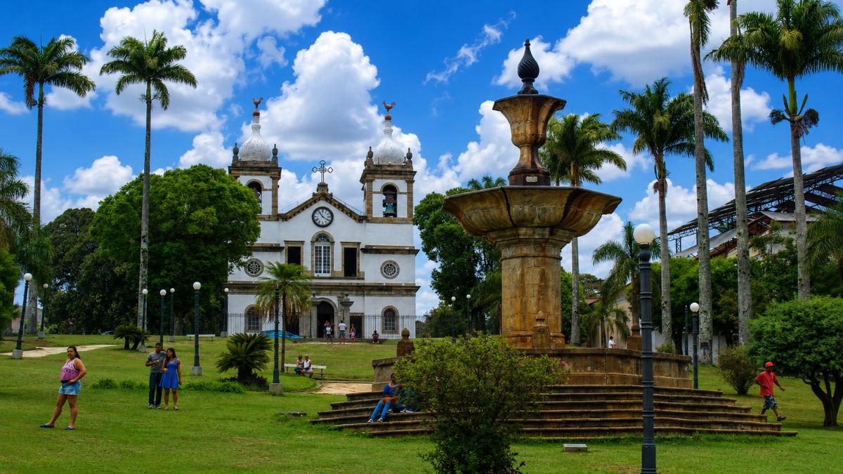 Fotografia em plano médio da Praça Barão de Campo Belo em Vassouras, exibindo um chafariz de pedra histórico em primeiro plano à direita. Ao fundo, centralizada na imagem, destaca-se a imponente Igreja Matriz de Nossa Senhora da Conceição com suas duas torres brancas sob um céu azul ensolarado e com nuvens. Há palmeiras imperiais altas distribuídas pelas laterais e gramados verdes com alguns pedestres caminhando e sentados.