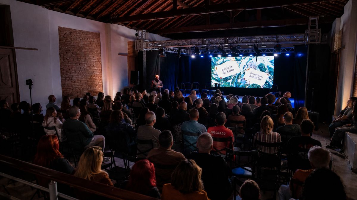 Fotografia em plano geral mostrando uma plateia sentada de costas em um auditório rústico com teto de madeira e paredes de tijolos aparentes. Ao fundo, há um palco iluminado com um homem em pé ao lado esquerdo e uma grande tela digital exibindo projeções gráficas coloridas no centro.