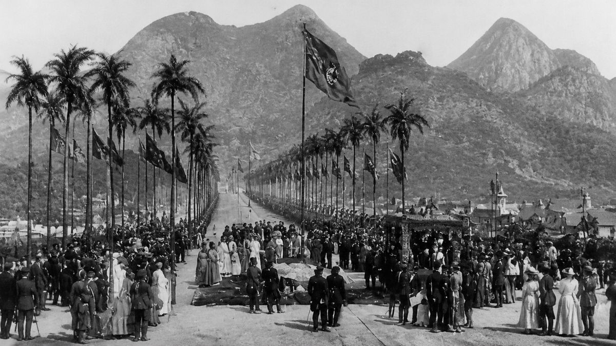 Fotografia antiga em preto e branco mostrando uma grande multidão reunida em uma avenida larga ladeada por palmeiras imperiais altas, bandeiras hasteadas e montanhas imponentes ao fundo durante um evento oficial.