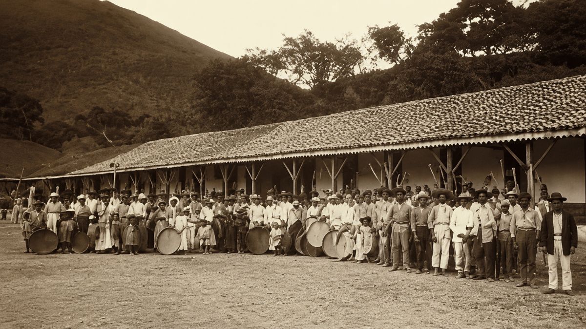 Fotografia em tom sépia mostra dezenas de trabalhadores enfileirados em frente a uma longa construção colonial com telhado de barro.